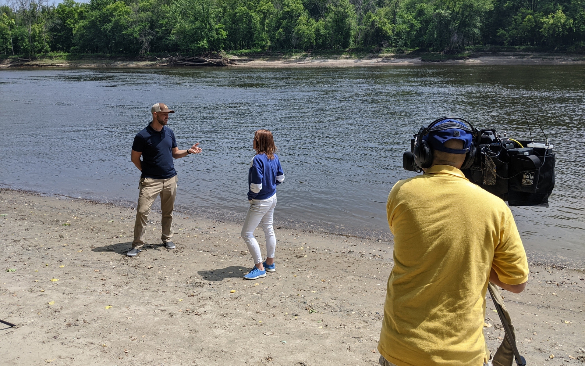 FMR Water Program Director Trevor Russell stands along the riverbanks, speaking to a journalist who is facing away from the camera. In the foreground, a camera operator in a yellow shirt films the two of them.