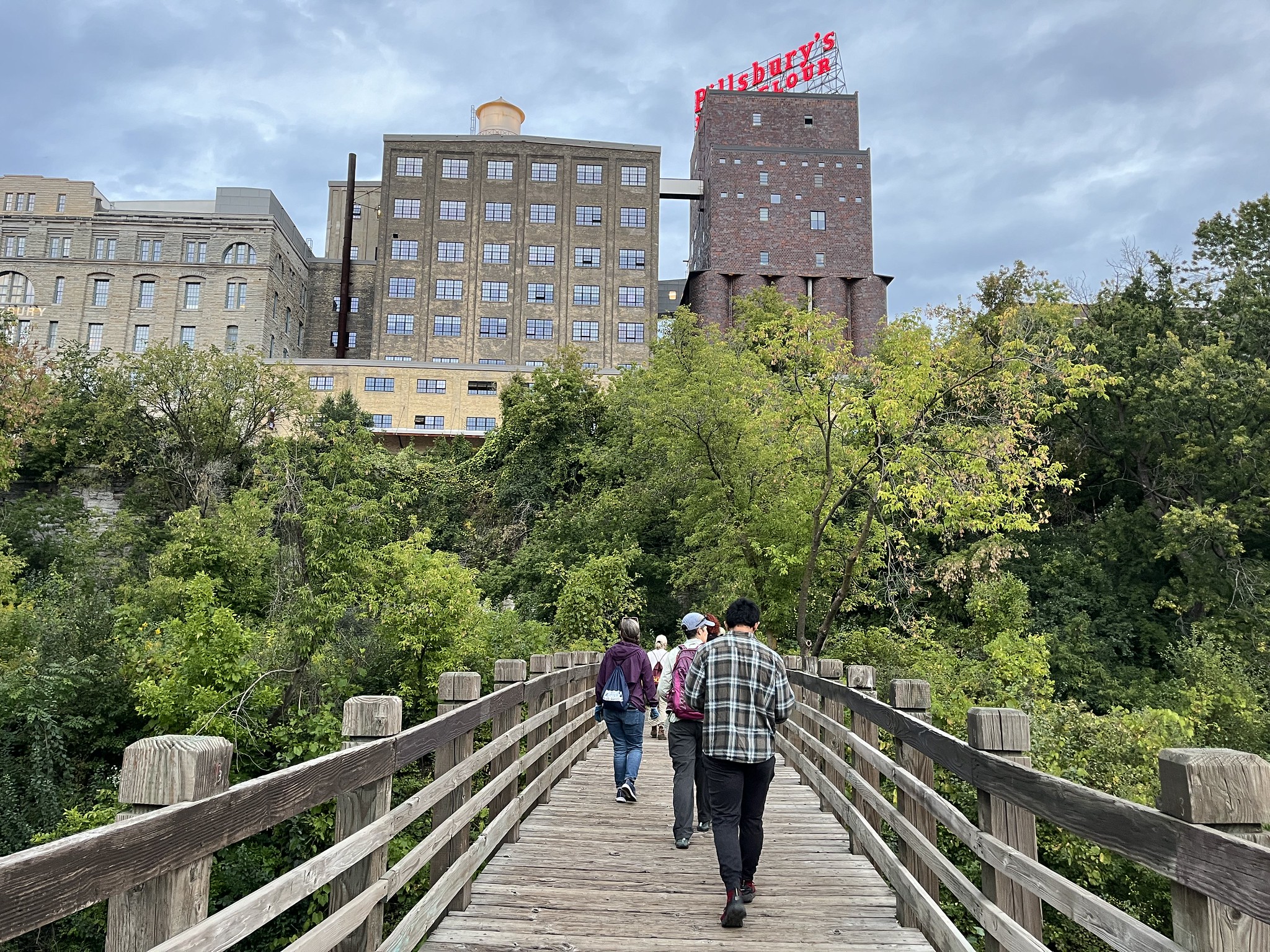 Volunteers walk across bridge toward forested area and Minneapolis buildings