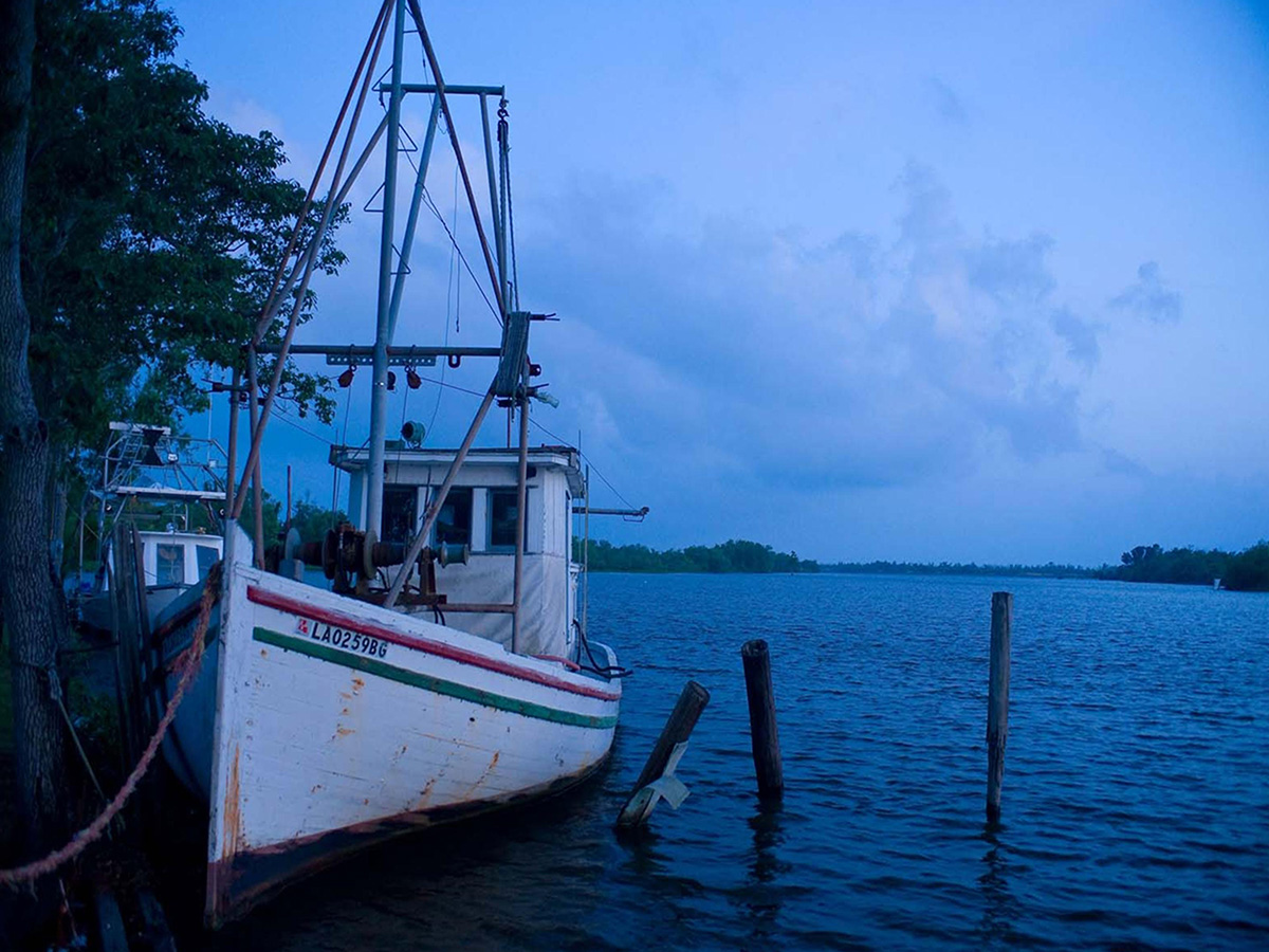 A white fishing boat along the shore of the Mississippi River in Louisiana.The water and sky are both a dark blue.