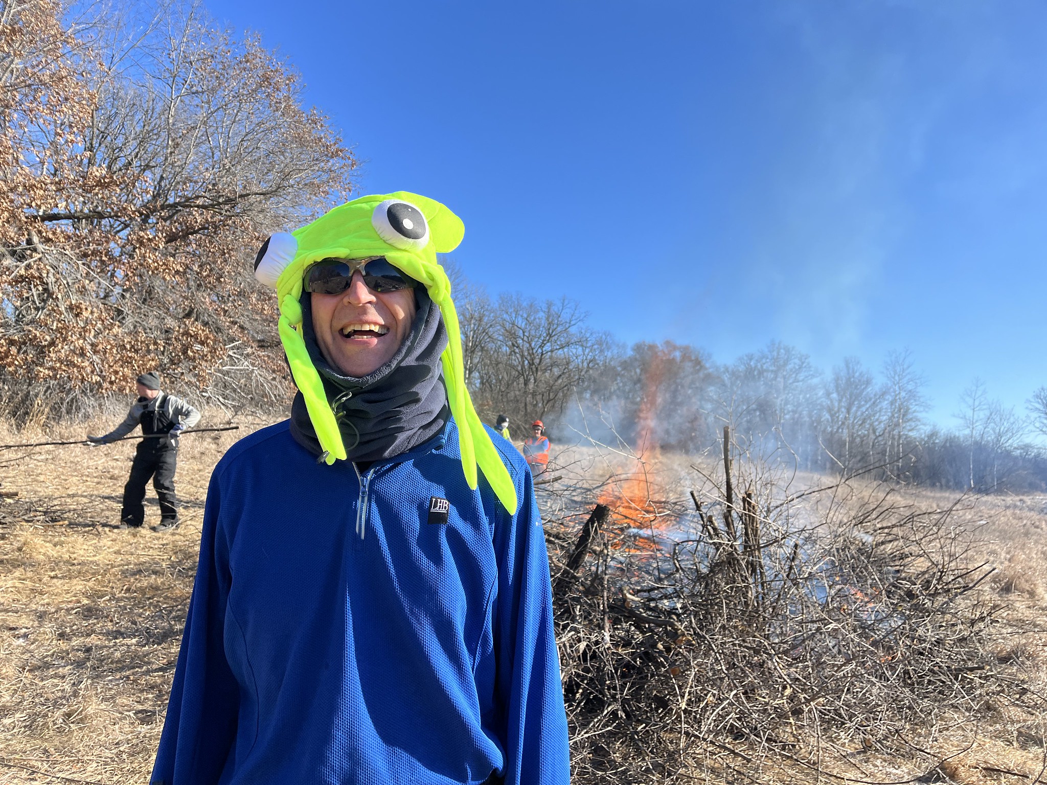 A volunteer in a bright green hat smiles in front of a buckthorn brush bonfire