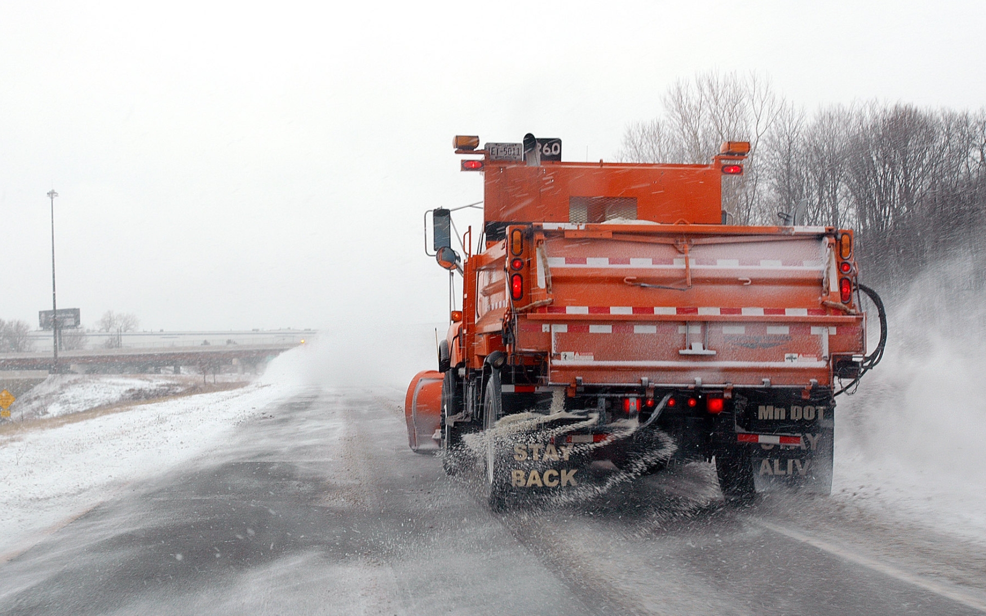 A red snow plow applying salt while traveling down a snowy road.