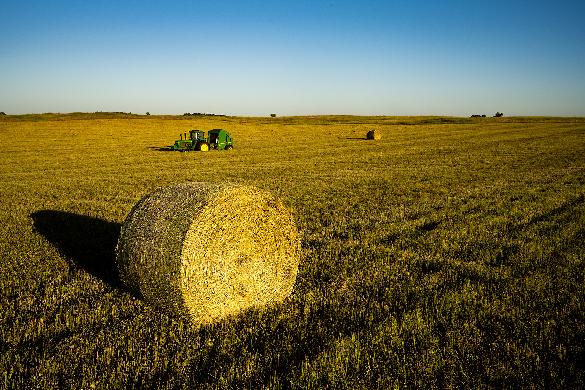 A bale of hay sits in the foreground. A baler can be seen in the distance.