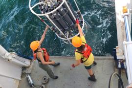 Two scientists stand at the edge of a boat, as a large piece of equipment is held at the surface of the water.