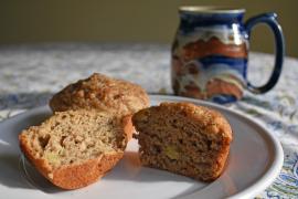 A recently baked cinnamon apple Kernza muffin is split apart on a plate. A mug can be seen on the table just behind it.