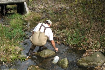 An MPCA staffer in waders reaches down to get a sample from shallow waters.