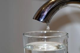 A drip of water falls from a metal tap, into a filling glass of water.