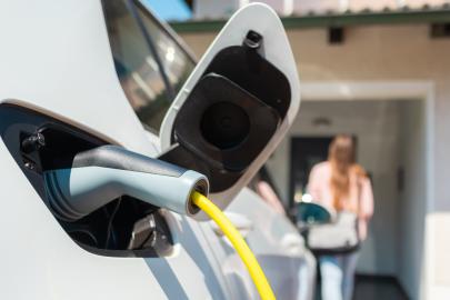 An electric vehicle plugged in and charging. In the background, a person is walking toward the front door of their home.