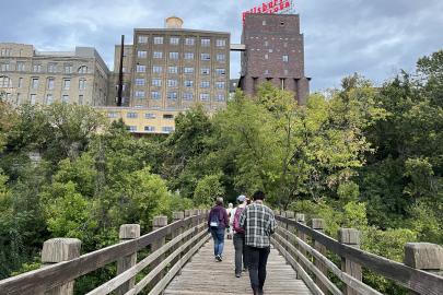 Volunteers walk across bridge toward forested area and Minneapolis buildings