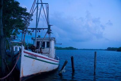 A white fishing boat along the shore of the Mississippi River in Louisiana.The water and sky are both a dark blue.