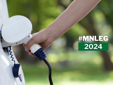 A hand puts a charging plug into an electric vehicle. Text says "MN Leg 2024"