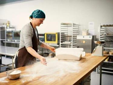 A baker at a large table, spreading flour as dough sits waiting to the side. 