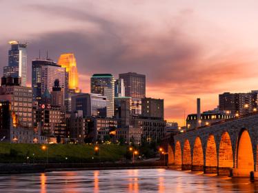 The Minneapolis skyline, reflective against the orange evening sky with the Mississippi River in the foreground.