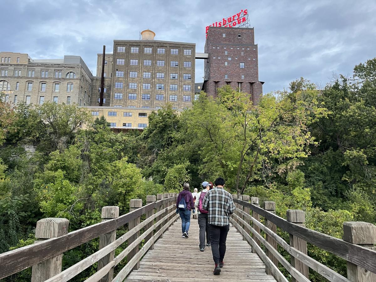 Volunteers walk across bridge toward forested area and Minneapolis buildings
