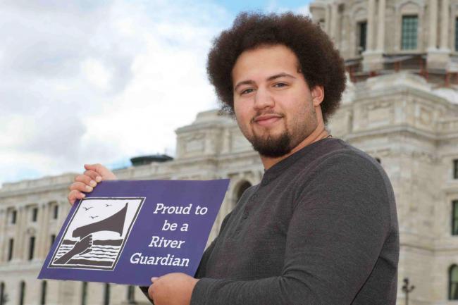A man stands in front of the MN state capitol with a sign reading "proud to be a river guardian"