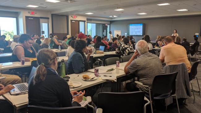 About 70 people sit at tables in a meeting room, facing a presenter.